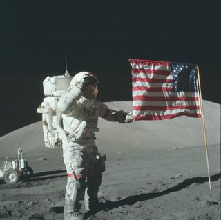 An astronaut in a bulky white spacesuit plants an American flag on the gray, dusty surface of the moon