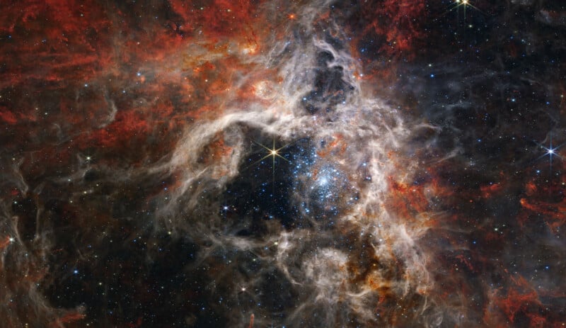 A stunning image of the Tarantula Nebula captured by the James Webb Space Telescope, showing a vivid and intricate tapestry of bright stars, swirling gas and dust clouds in hues of red, orange and black, set against a dense star-studded background.