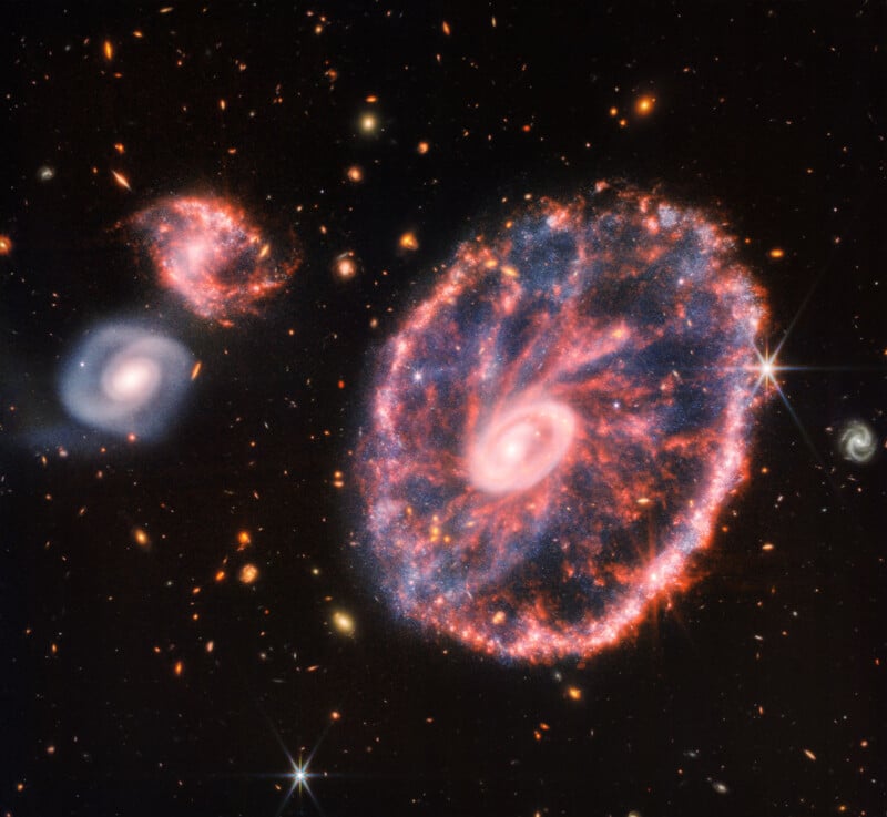 An image of the Cartwheel Galaxy taken by the James Webb Space Telescope shows a bright ring-like structure with a vibrant, luminous core. Spiral galaxies and other celestial objects surround it, against the dark backdrop of star-studded space.