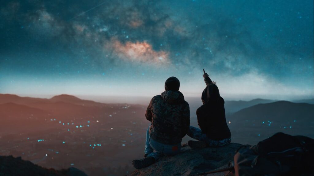 two people sitting down and looking at the night sky with the naked eye, the person on the right is pointing up at the stars.