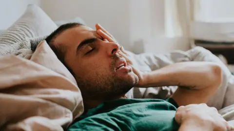 Getty Images A man looking sick and holding his head in bed