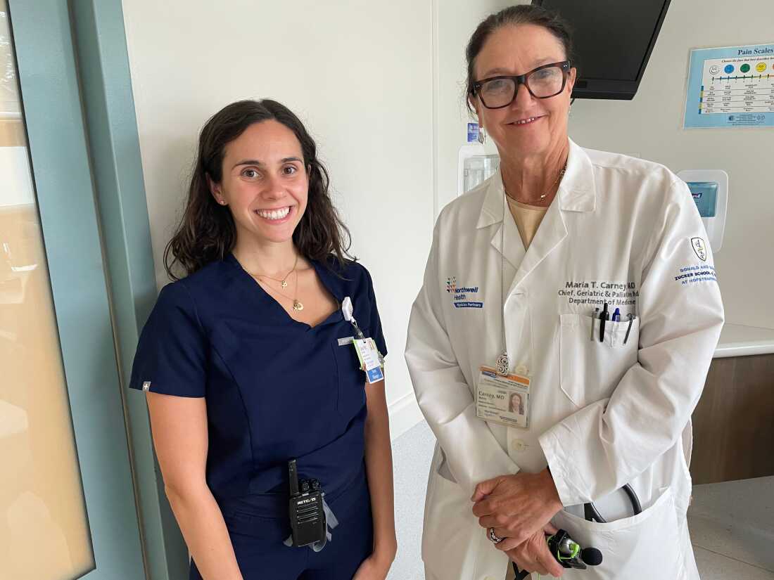 Geriatrician Maria Carney (right) wears a white coat and glasses and stands next to head nurse Cara Gibbons, who wears a dark blue hospital scrub. They are in a hospital room.