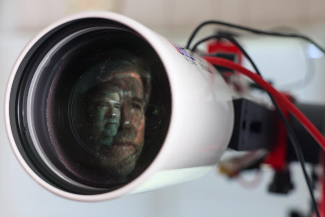 A bearded man's face is reflected in the lens of a telescope.