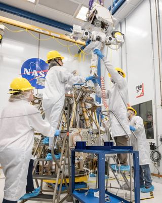 Four technicians in white coats and yellow helmets assemble a robotic explorer in a clean room.