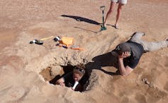 A scientist sits in a small hole to examine and sample the sediment, while his colleagues look on.