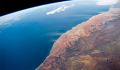 Satellite image showing the blue of the South Atlantic on the left, then the Namib Sand Sea, with its distinctive orange-red sand, then inland to the Great Escarpment Mountains. Dust plumes blown from the dried-up river valleys are visible.