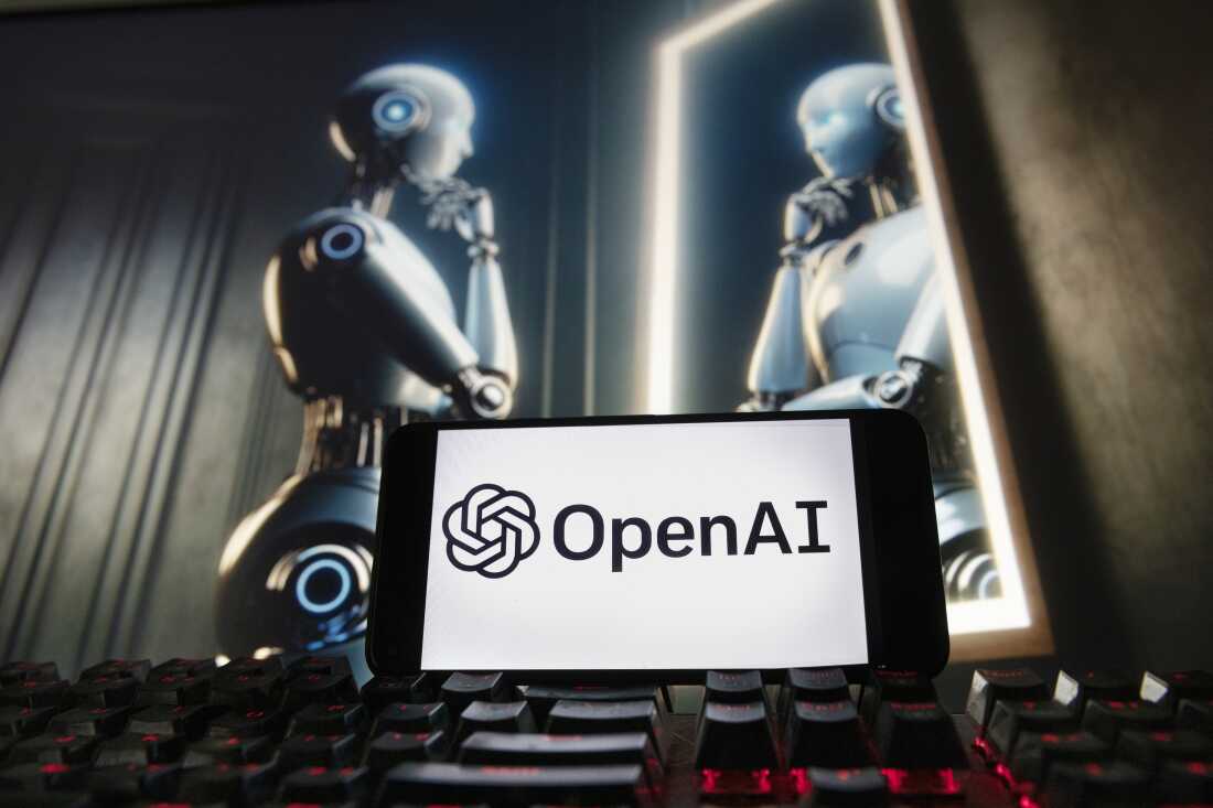 In the foreground, a black keyboard with red backlighting. Behind it, a tablet in landscape orientation displays the name OpenAI and the technology company's logo on its screen. In the background, a metal robot resembling a human looks at itself in a mirror.