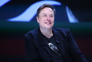 a man wearing a black blazer over a black t-shirt smiles on stage at a press conference