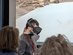 A man wearing a black virtual reality headset over his face walks in front of a large screen with a rocky shoreline, other people watching in the foreground