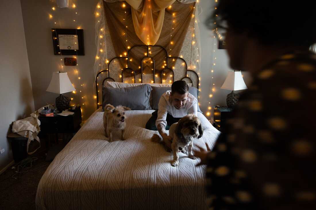 Thomas Guzowski greets his dogs — a 13-year-old shih tzu and a 15-year-old poodle-terrier mix — as they climb onto the bed on July 18 in Redlands, California. The bed has a white bedspread and two blue pillows. String lights hang on the wall behind the bed.