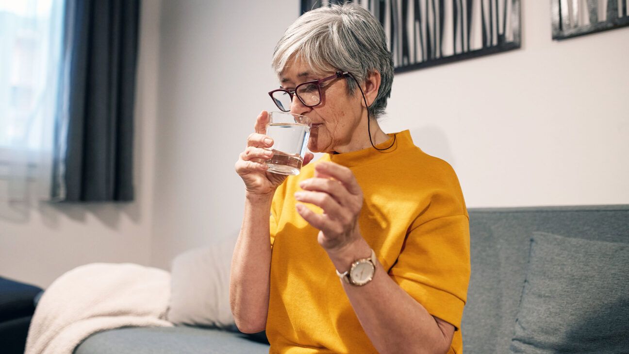 A woman wearing glasses taking a supplement with a glass of water.