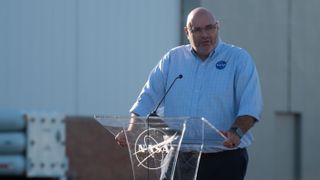 A bald man wearing a collared shirt with the NASA logo stands at a clear podium with a NASA logo, in front of a hangar in the background.