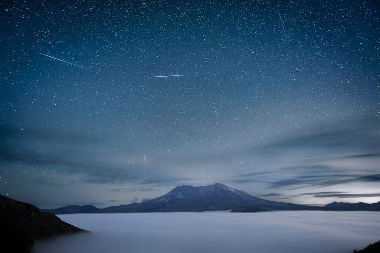 A misty mountain in the distance with a misty lake in the foreground and several light trails in the sky.