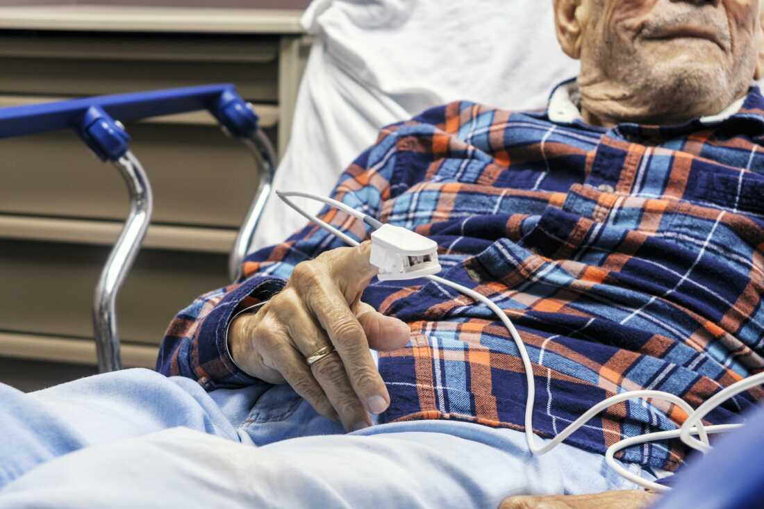 An elderly man lies in a hospital bed with safety rails. He is wearing a blue and red plaid shirt and his pulse oxygen level is measured with a device attached to his finger. His face is only partially visible. 