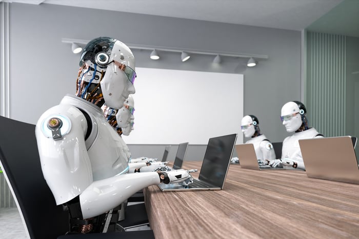 Several humanoid robots type on laptops while sitting at a long conference room table. 