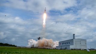 A triple-core rocket takes off under a sunny, partly cloudy blue sky. Smoke billows below, next to a hidden launch tower, near a long white hangar with SPACEX written on it. To the left, a bird takes flight.