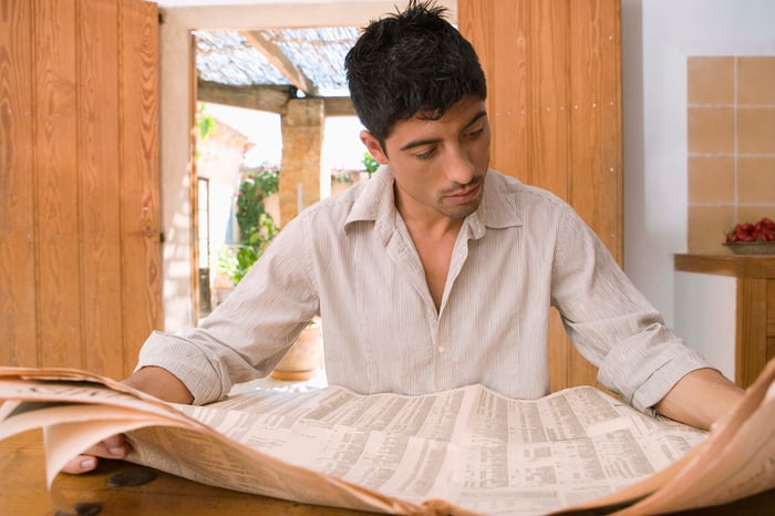 A person reading a financial newspaper sitting at a table at home.