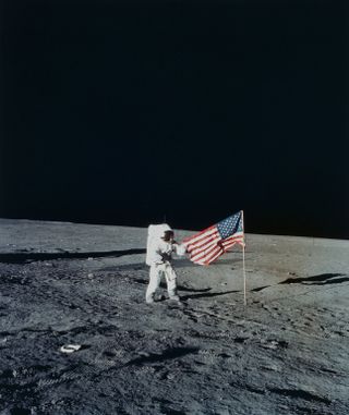 An astronaut in a bulky white spacesuit plants an American flag on the gray, dusty surface of the moon